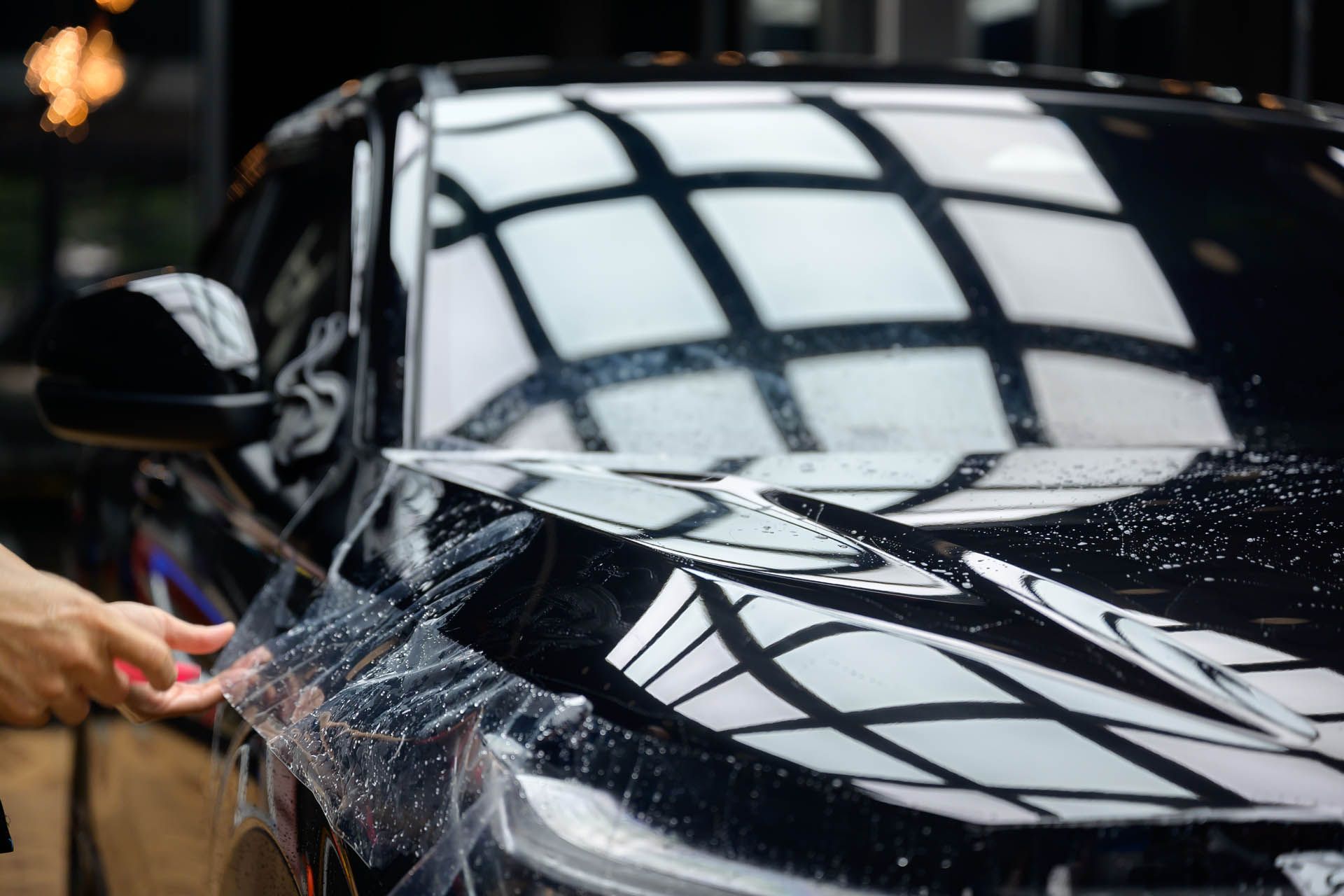 A hand carefully applies a clear protective film to the glossy black hood of a car.
