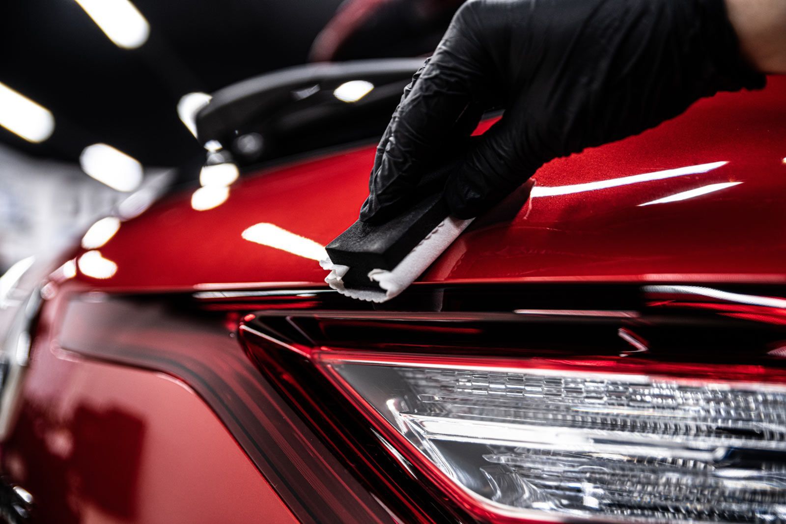 A gloved hand applies a ceramic coating to a glossy red car exterior using a foam applicator block.