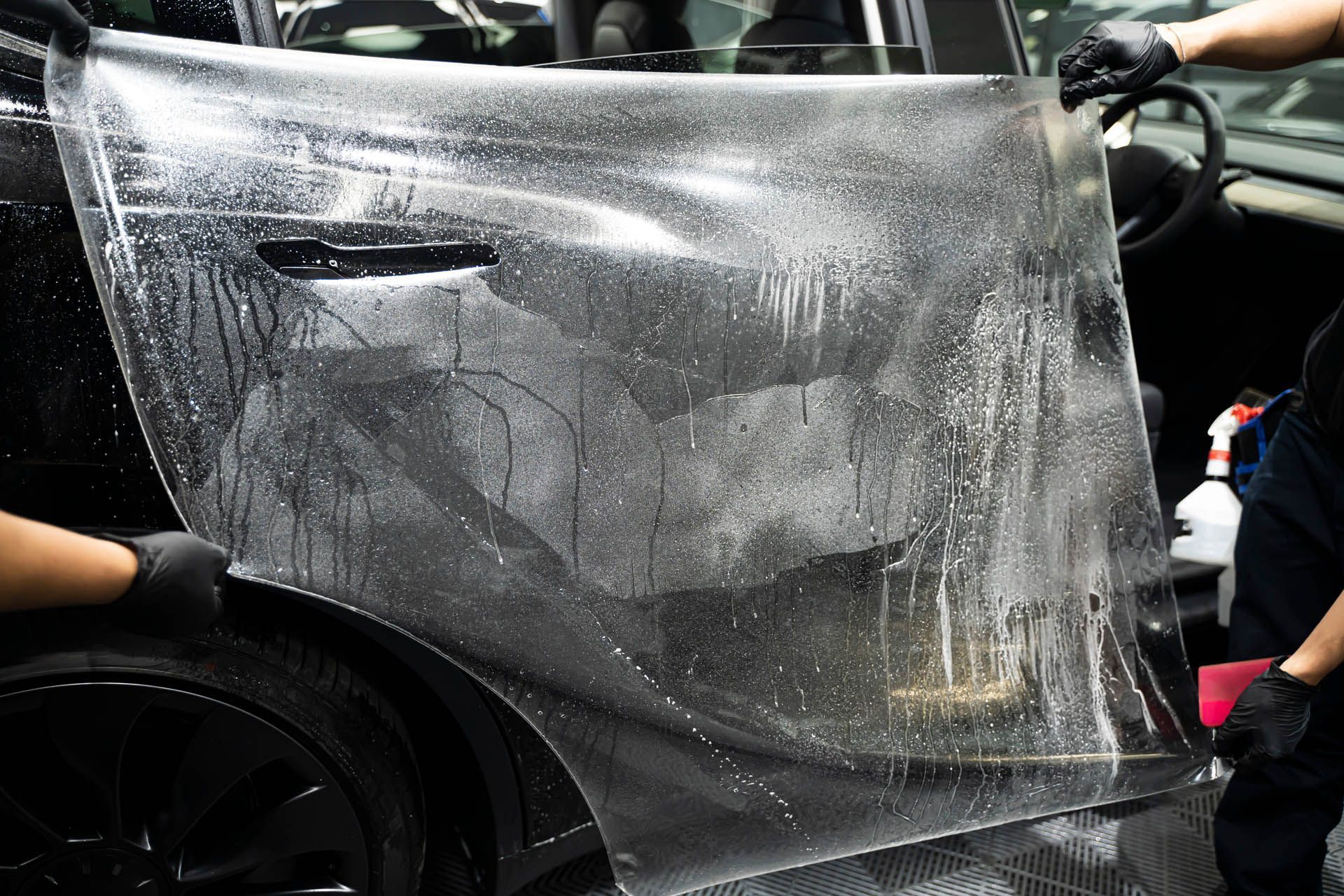 Two technicians in black gloves apply a clear protective film to the side of a black car.