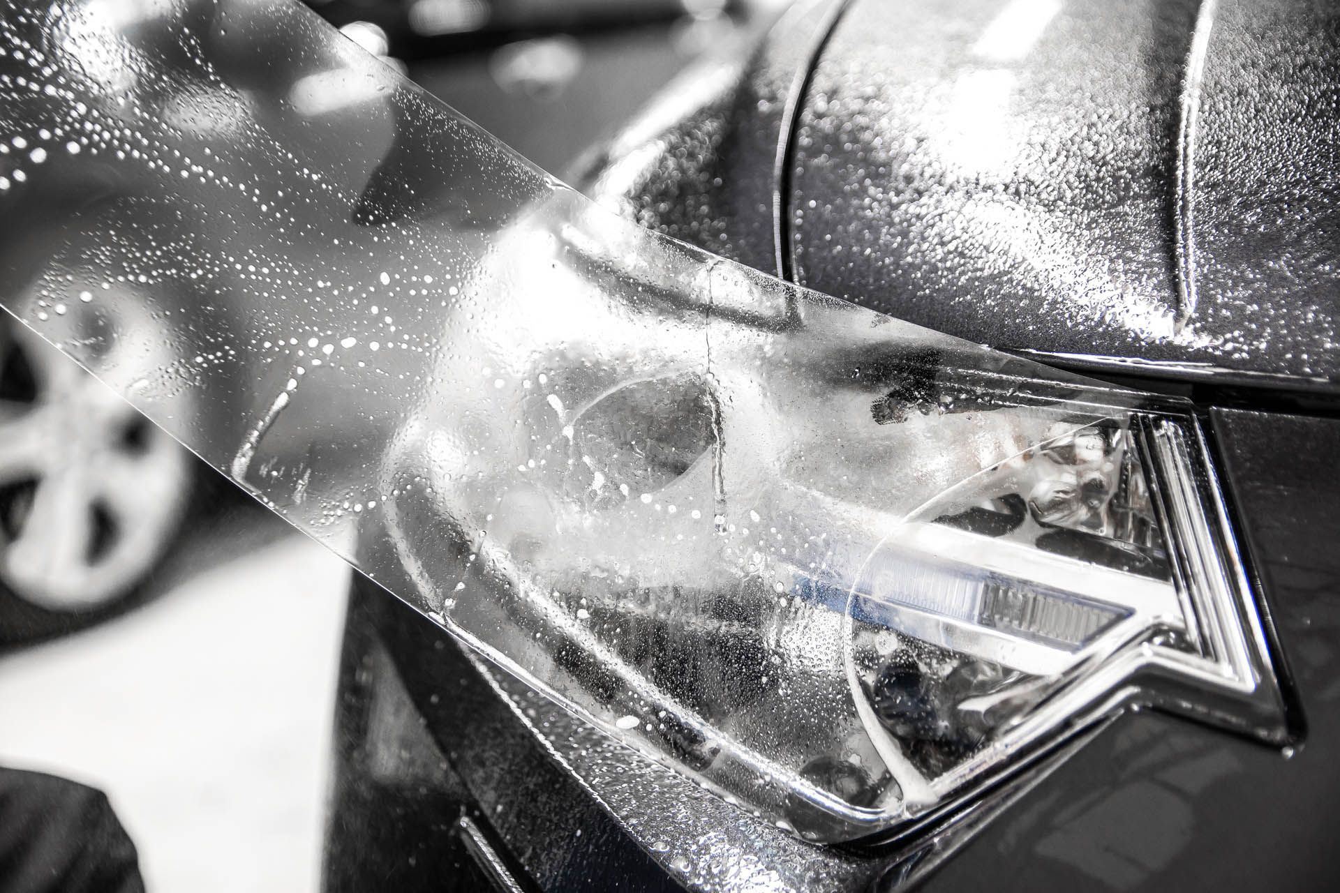 A hand applies a protective clear film to a car’s headlight, showing water droplets between the plastic and the light.