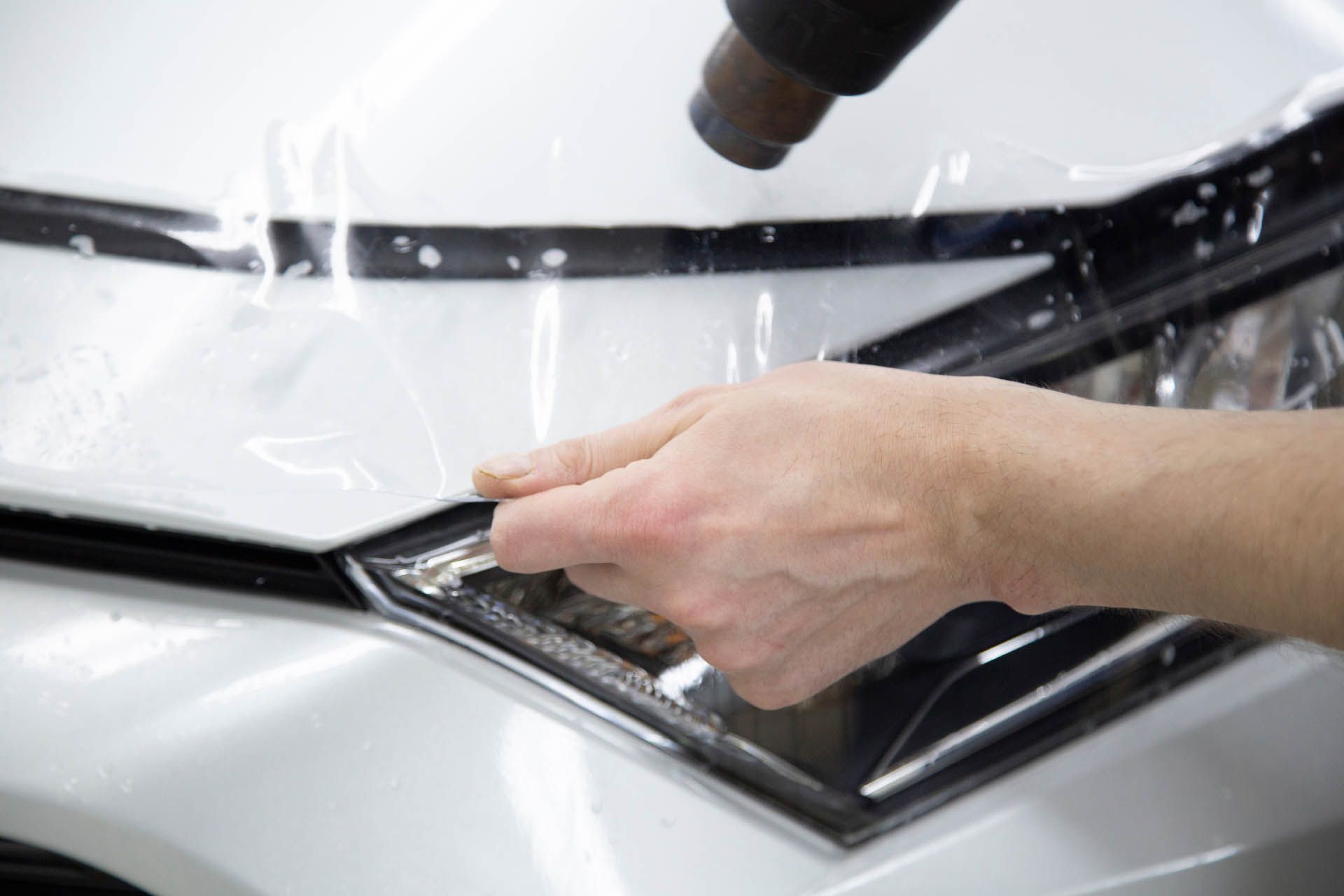 A hand uses a heat gun to apply a protective clear film over the white hood and headlight of a vehicle.