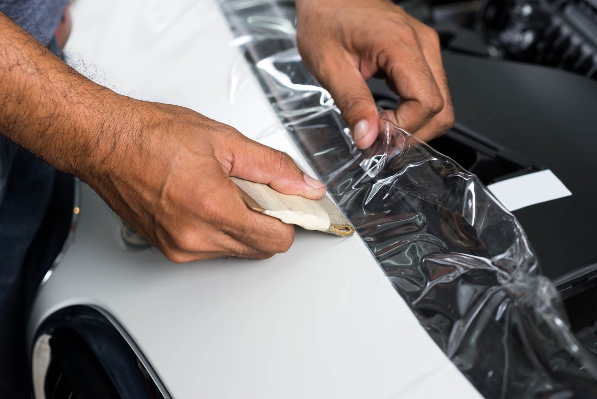 Hands use a squeegee to smooth a clear protective film onto the white hood of a car.