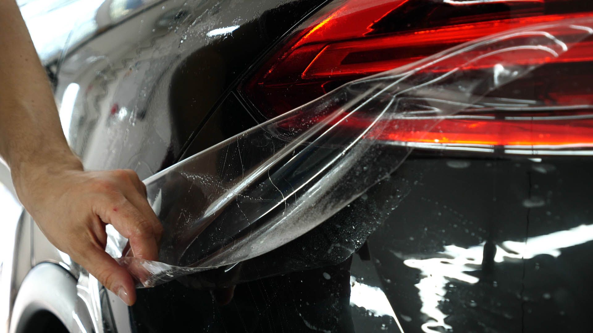 A person applies clear protective film over a dark car's rear panel near the tail light.