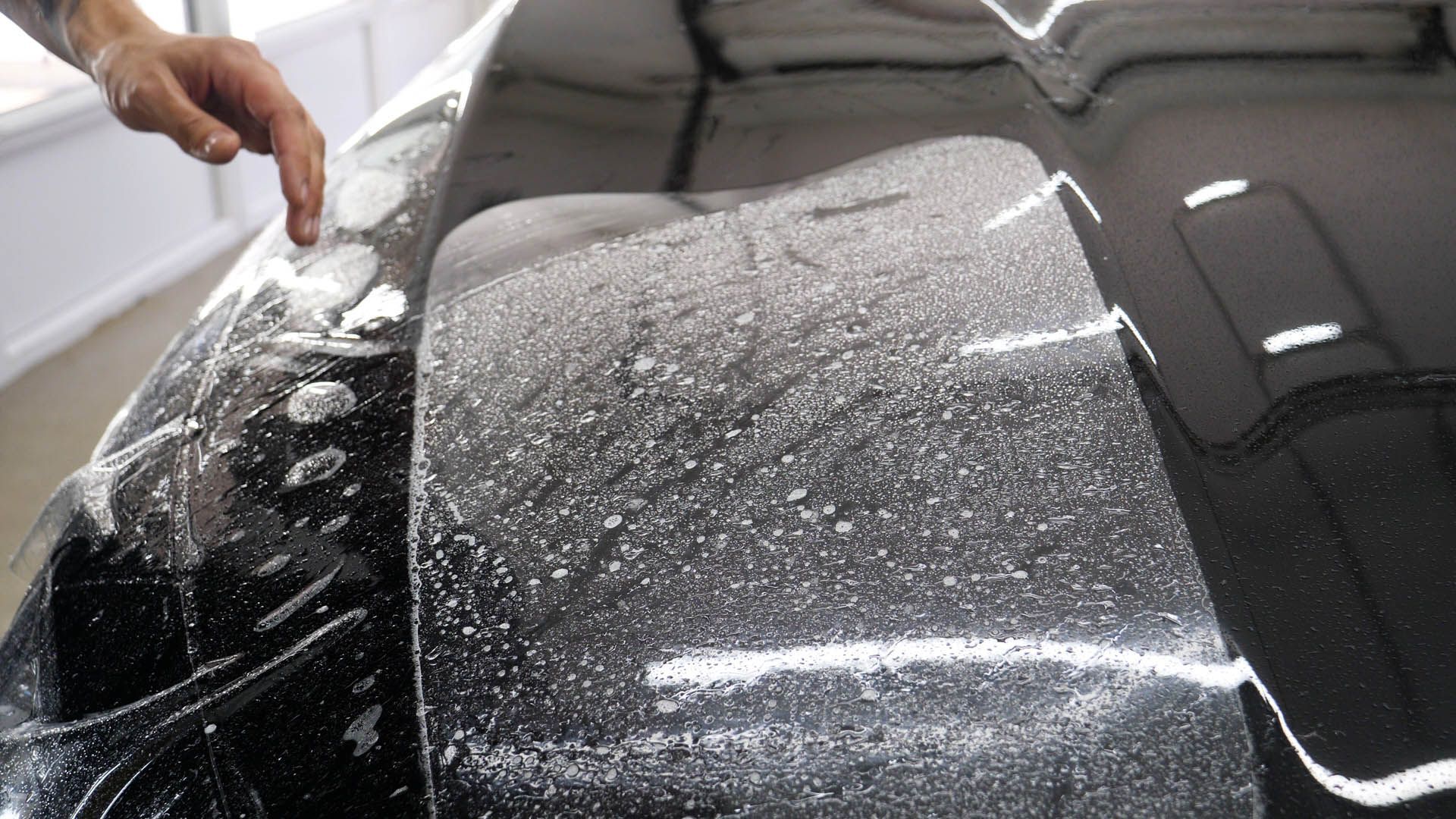 A hand applying a protective clear film to a black car hood in a workshop.