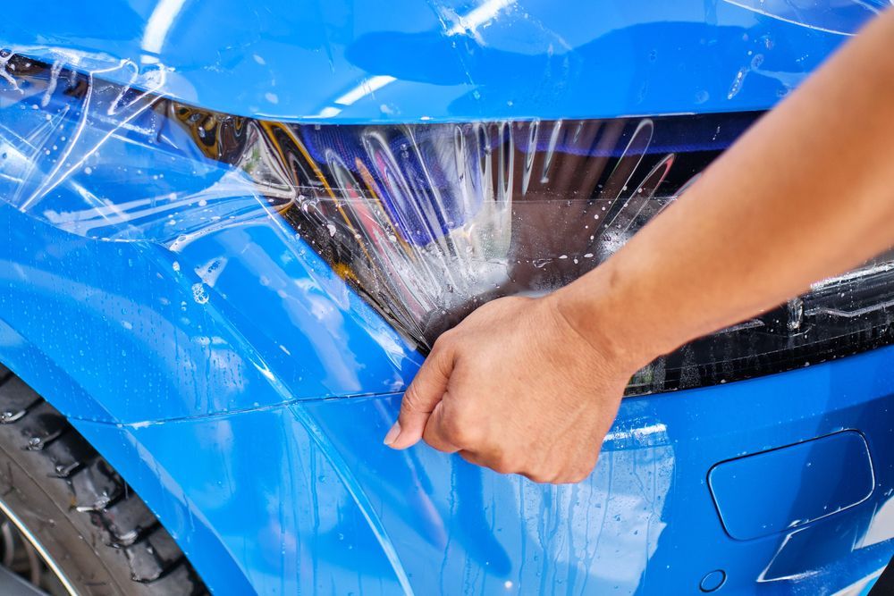A technician applies a clear protective film to the headlight and bumper of a bright blue car.