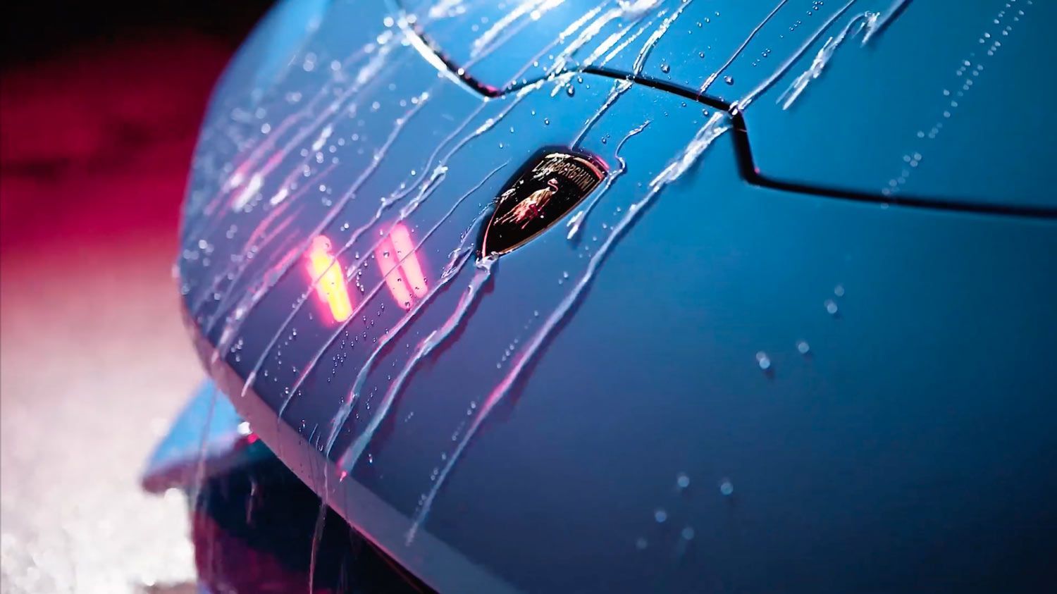 Close-up of a blue Lamborghini hood with water streaming down over the brand logo under red and blue ambient lighting.