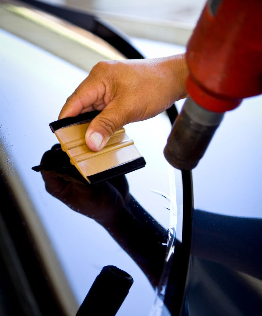 Squeegee and heat gun pressing window tint film against the edge of a dark car window during installation.